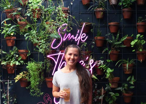 A dark haired woman standing in front of a wall of green plants and a neon sign that reads 'SMILE'. She's holding a mocktail.