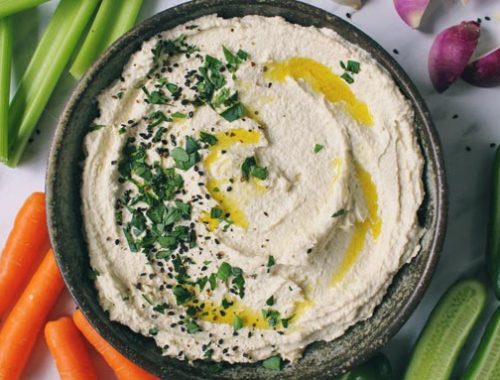 A bowl filled with bean-free hummus, sprinkled with chopped parsley and colorful vegetables are laid out around bowl