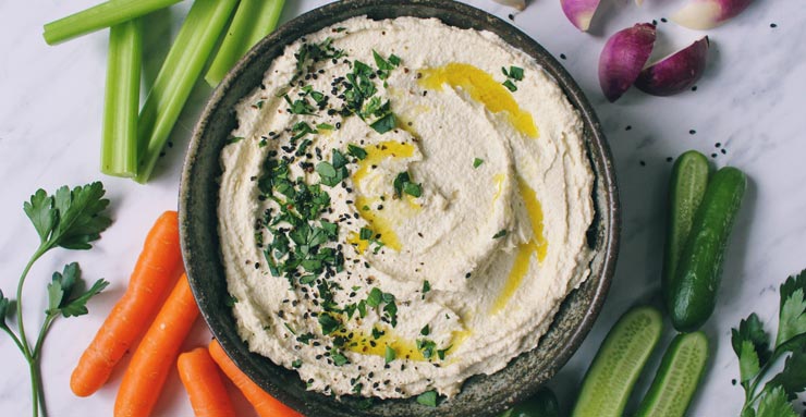 A bowl filled with bean-free hummus, sprinkled with chopped parsley and colorful vegetables are laid out around bowl